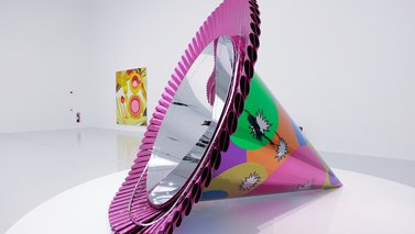 A large, multicoloured stainless steel sculpture of a party hat sitting on its side in a gallery