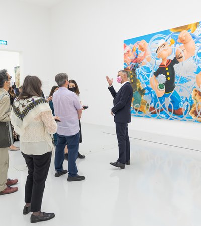 Massimiliano Gioni talking to a group of visitors in a gallery, standing in front of Jeff Koons' painting Triple Popeye