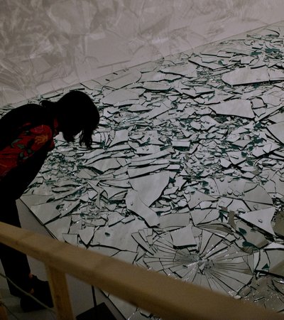 A woman standing in a gallery overlooking a large floor filled with pieces of broken mirrored glass