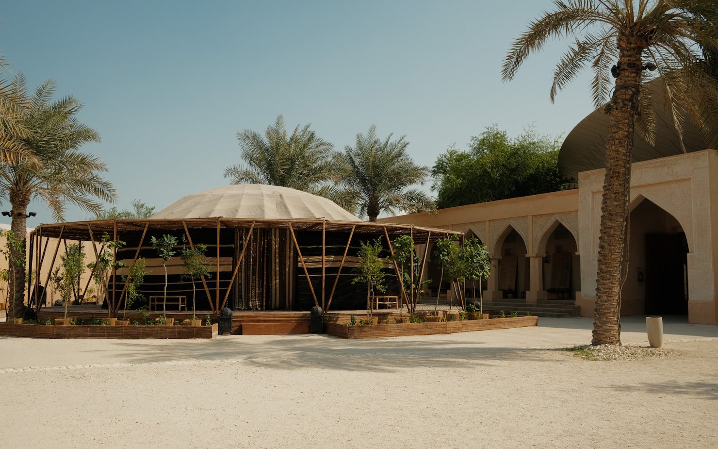A tent-like structure with bamboo poles holding the perimeter sits in the middle of a courtyard flanked by palm trees.