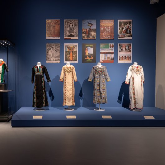 A gallery showcasing Palestinian dresses embroidered with a symbol of the Palestinian flag with posters in the back depicting war times in Palestine
