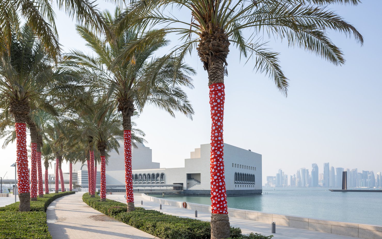 Palm trees wrapped in red and white polka dot material line a path on the water leading up to a white building.
