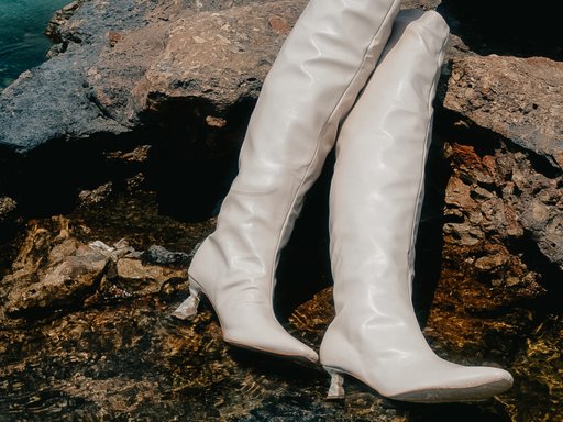 A pair of high-heeled white leather women's boots positioned on rocks by the sea.