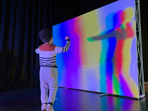 A boy standing in front of a white screen reflecting his shadow in various colour