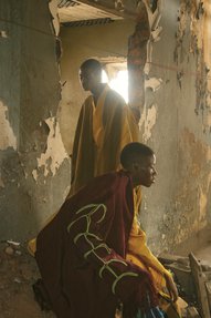 Two young men, dressed in loose robes stand inside a room within a derelict building.