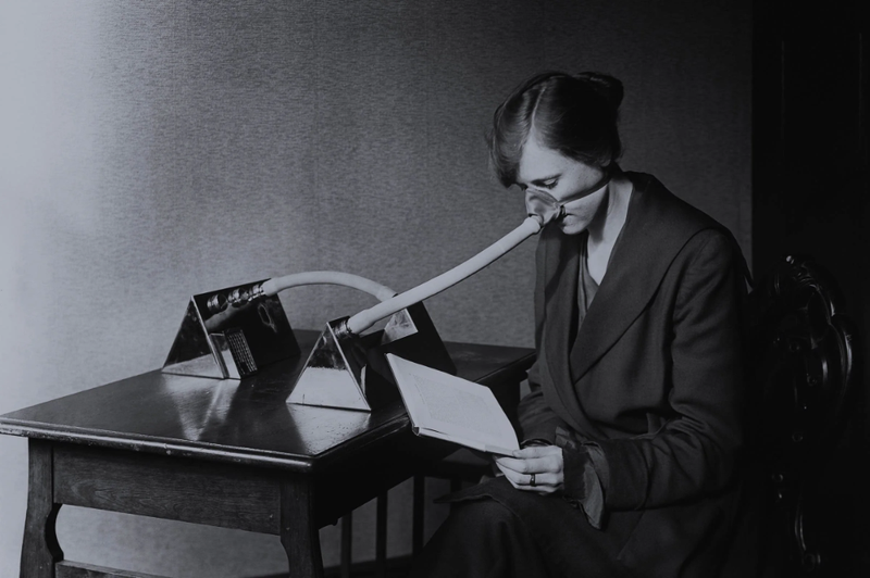 Woman wearing a respirator connected to rubber and metal seated at a desk, holding a book. Picture is from the earlier 20th century.