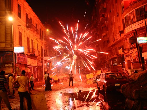 A city street at night, crowd of people and a bright explosive firework lighting the night sky.