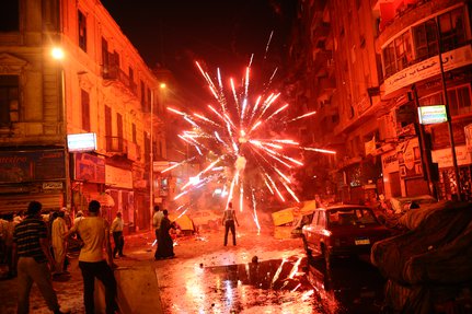 A city street at night, crowd of people and a bright explosive firework lighting the night sky.