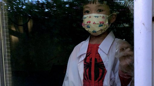 A little girl looking out behind a window glass wearing a spiderman costume with a doctor's gown and a mask on her face