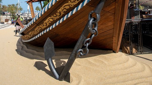 A view of the front of a large wooden boat (dhow) with a white sail, painted decorative sides and huge metal anchor standing on the sand
