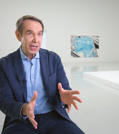 Jeff Koons at his exhibition, behind him are his public art displays of a orange balloon dog on the left and a play dough on the right