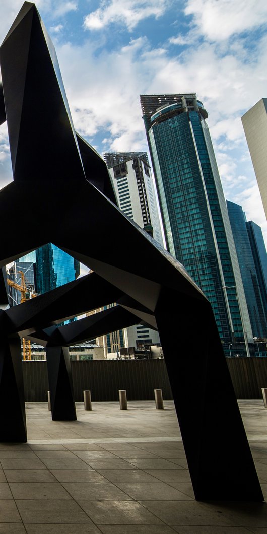 A view of Smoke, a black metal geometric sculpture by Tony Smith set against a backdrop of tall buildings in Doha