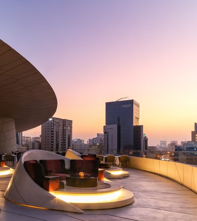 A view of the rooftop terrace seating at the Jiwan retaurant, National Museum of Qatar lit up at night with of Doha in the background